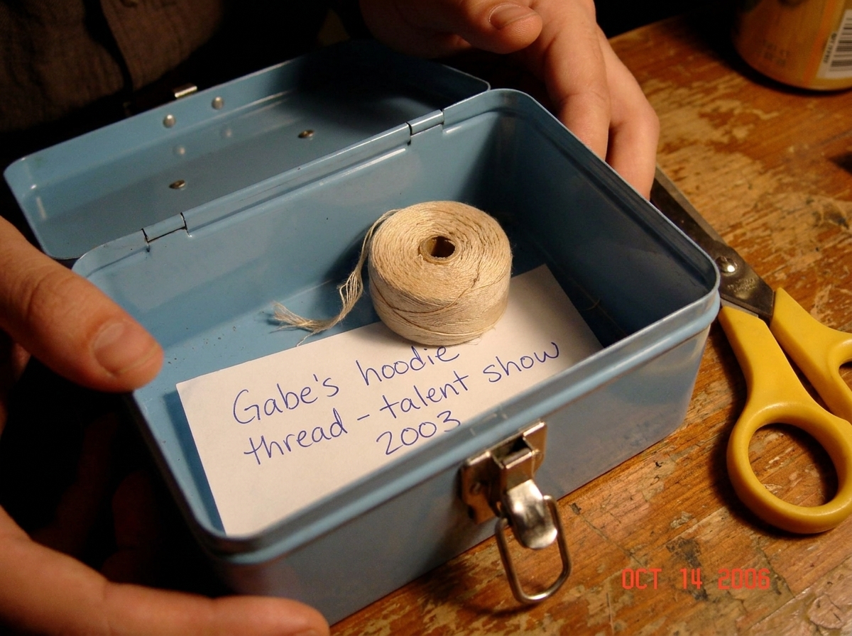 A close-up photo of a light blue sewing box opened to reveal a small, faded, light-colored thread coiled inside. The thread is slightly frayed at one end. The photo is taken in warm indoor lighting on a wooden table. A pair of yellow-handled sewing scissors sits next to the box. A handwritten note i