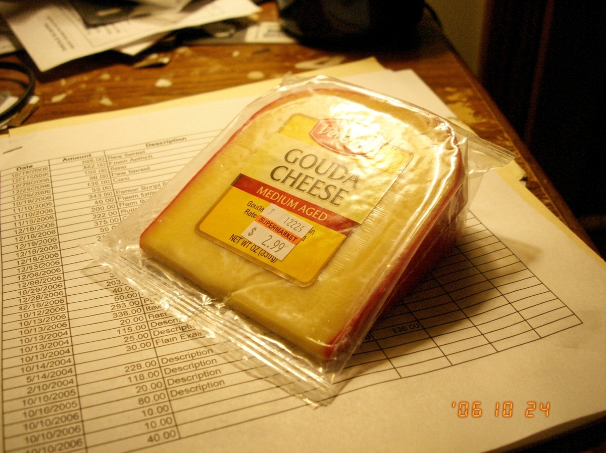 A close-up of a piece of gouda cheese in its wrapper, sitting on top of a printed spreadsheet. The focus is soft and the lighting is warm and golden, creating a halo effect around the cheese.