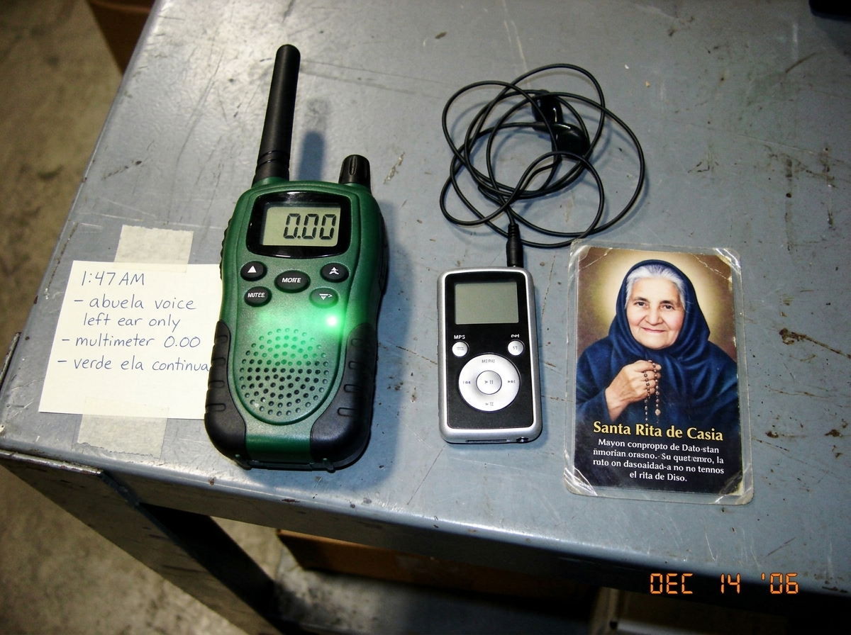 A photo of a green-glow walkie talkie sitting next to an MP3 player and a small Catholic prayer card of a grandmother, all arranged on a metal warehouse table. The walkie shows 0.00 on its display but emits a faint green glow from its speaker grille. A handwritten note beside it reads '1:47 AM - abu
