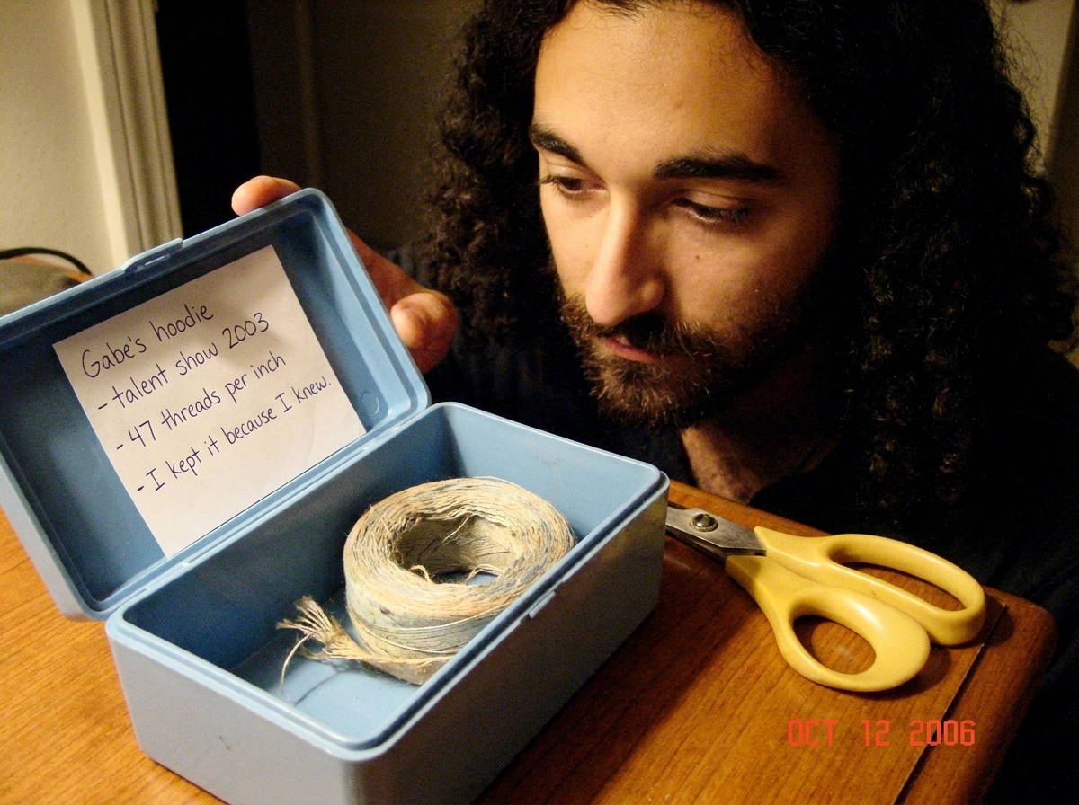A close-up photo of a light blue sewing box opened to reveal a small, faded, light-colored thread coiled inside. The thread is slightly frayed at one end. The photo is taken in warm indoor lighting on a wooden table. A pair of yellow-handled sewing scissors sits next to the box. A handwritten note i