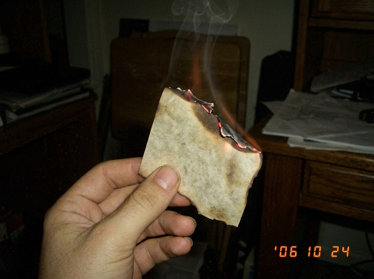 A close-up of a hand holding a piece of burnt vellum paper, smoke curling upward in a dim room, the edges of the paper glowing orange