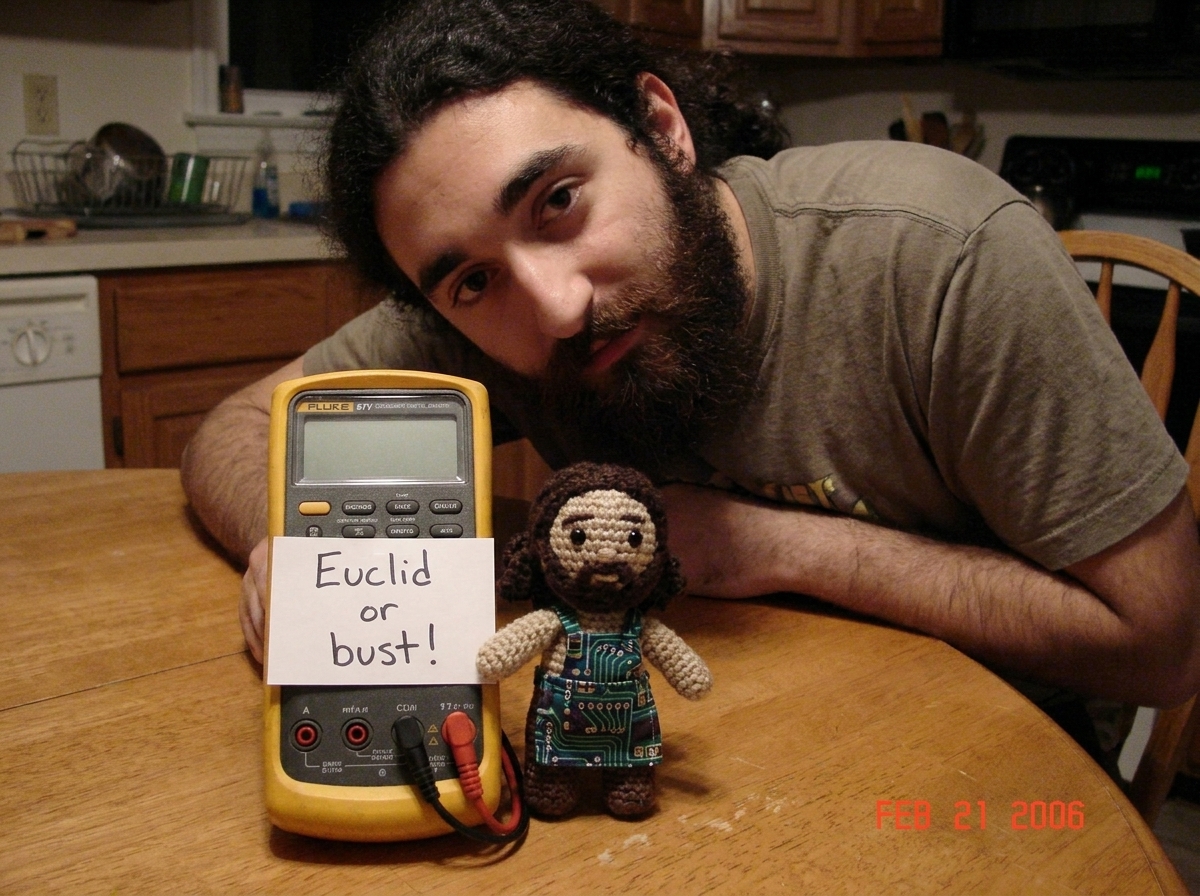 A photo of a Fluke 87V multimeter sitting on a kitchen table next to a crocheted Gabe-able doll wearing a tiny circuit-board apron. A handwritten note taped to the meter reads 'Euclid or bust!' The multimeter display is blank, showing no readings.
