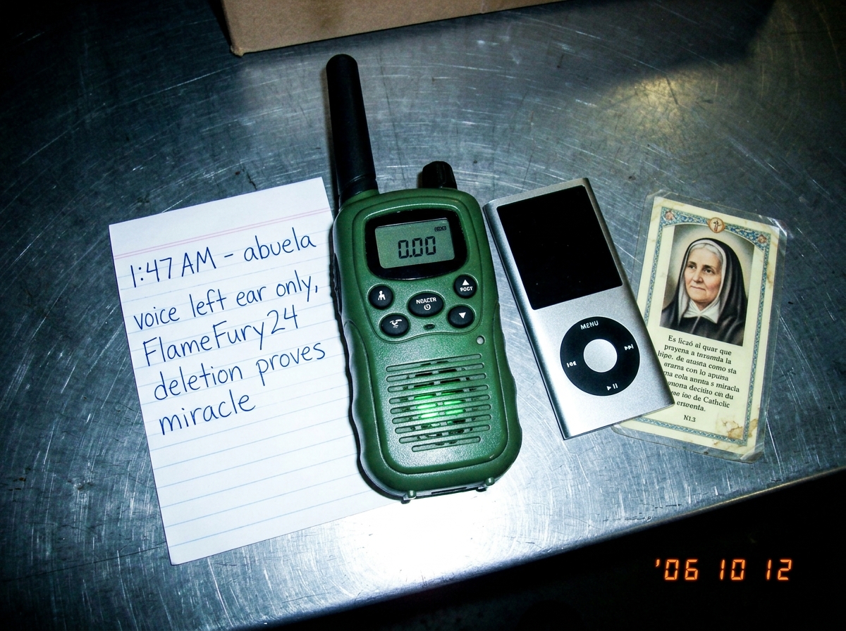 A photo of a green-glow walkie talkie sitting next to an MP3 player and a small Catholic prayer card of a grandmother, all arranged on a metal warehouse table. The walkie shows 0.00 on its display but emits a faint green glow from its speaker grille. A handwritten note beside it reads '1:47 AM - abu