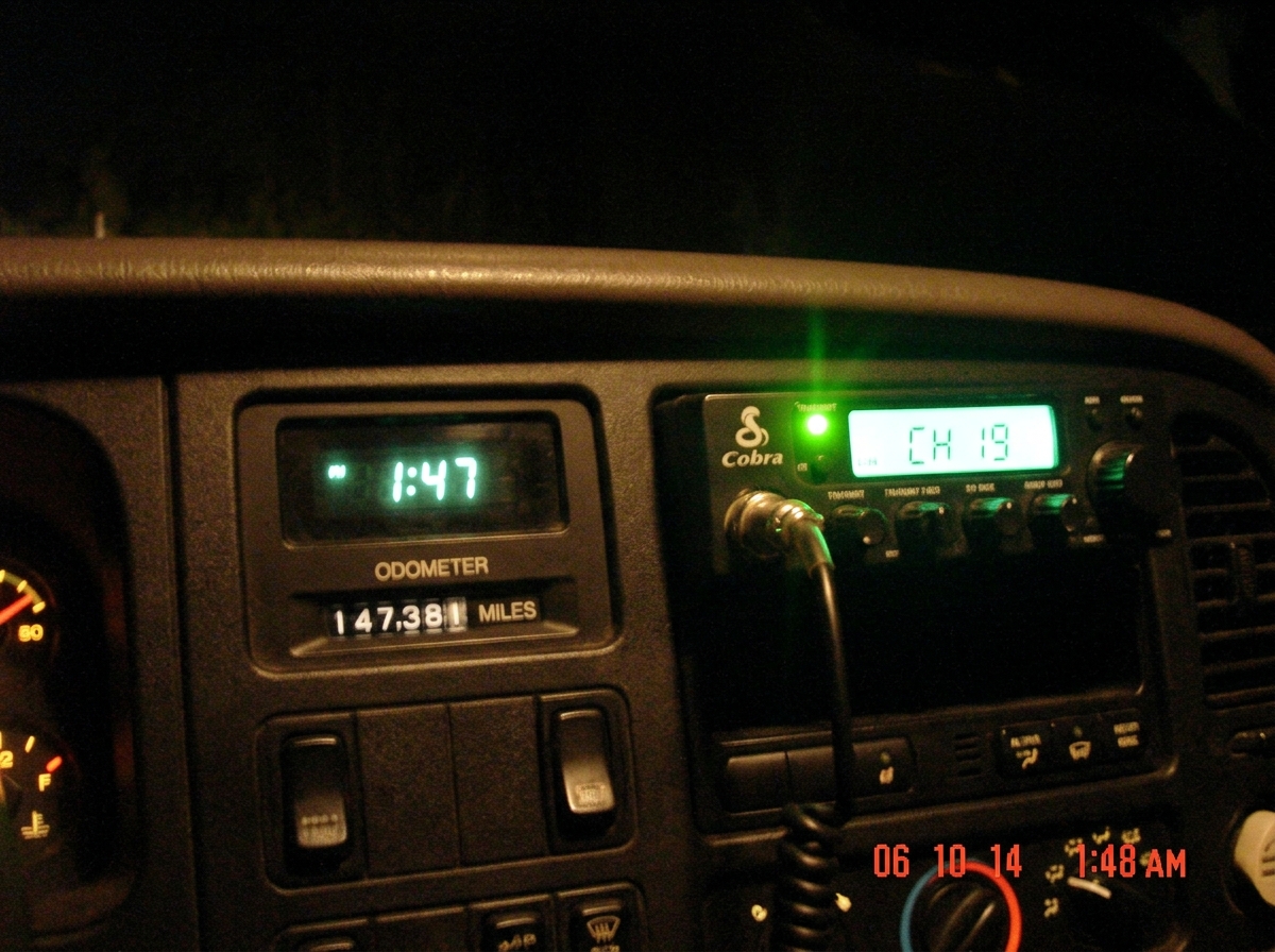 A close-up photo taken from a truck cab at night. The dashboard clock reads 1:47 in green digital numbers. The CB radio next to it has its green transmit light glowing. Below, the odometer reads 147,xxx miles. The photo is slightly blurry and lit only by the dash lights.