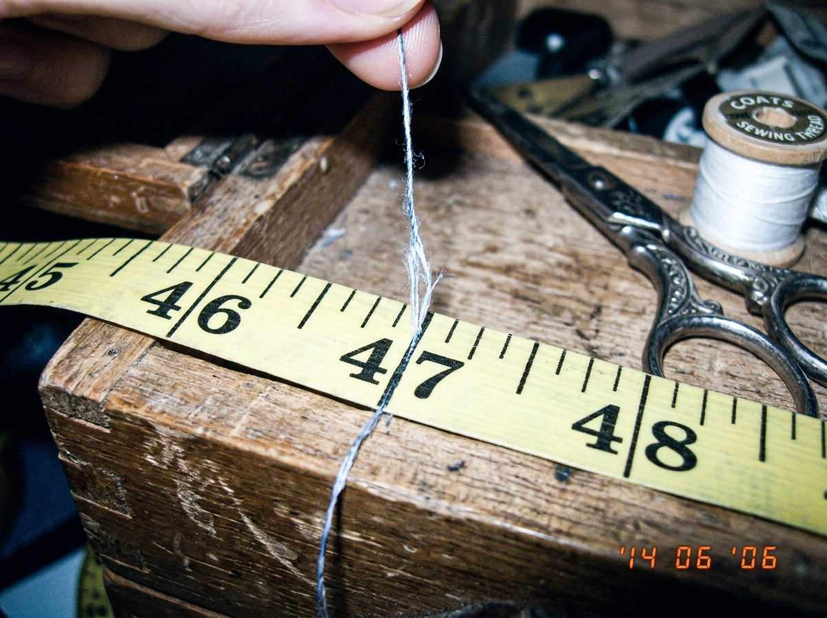 A close-up photo of a single frayed cotton thread held against a tailors measuring tape, stretched across a wooden sewing box. The thread lines up exactly with the 47-inch mark on the tape. In the background, a pair of vintage silver sewing scissors and a spool of white thread are visible. The photo