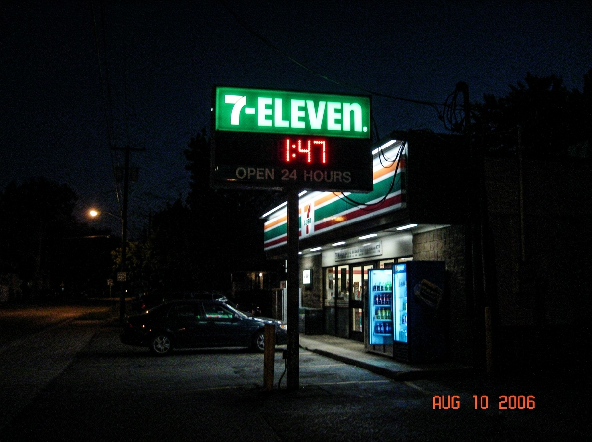 A grainy nighttime photo of a 7-Eleven store sign glowing green in the dark. The clock on the sign reads 1:47. Below it, a cooler door is slightly ajar with a faint blue light leaking out. The image has a date stamp 'Aug 10 2006' in the corner and looks like it was taken with a cheap flip phone came