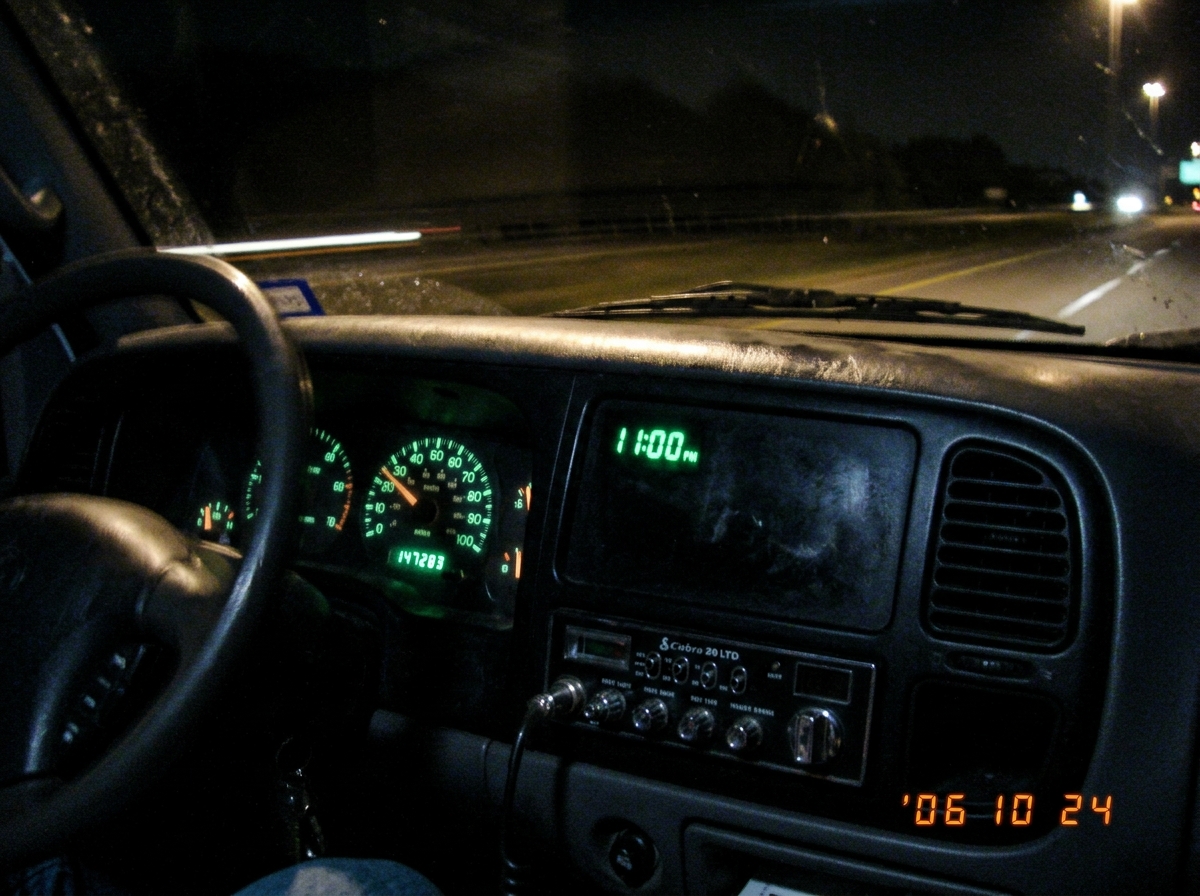 A blurry photo taken inside a truck cab at night. The dashboard clock shows 11:00 PM in green digital numbers. The CB radio below is dark - no lights on. The odometer reads 147,2xx miles. The photo is lit only by the dim green dash lights.