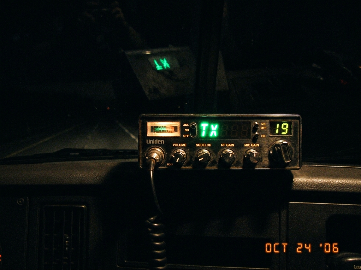 A photo taken from a truck cab at night. The green transmit light on a CB radio is glowing in the darkness. The dashboard clock is not visible - only the CB face with its dials and the bright green indicator light, casting a faint reflection on the windshield. No other lights in the cab are on.