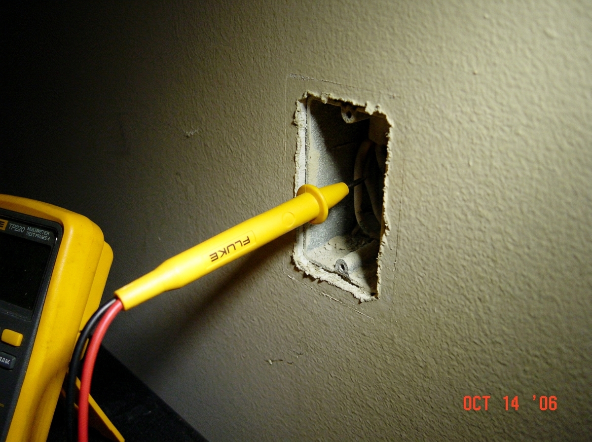 A close-up photo of a yellow Fluke multimeter probe pointing at a blank, empty wall where a socket should be. The lighting is dim.