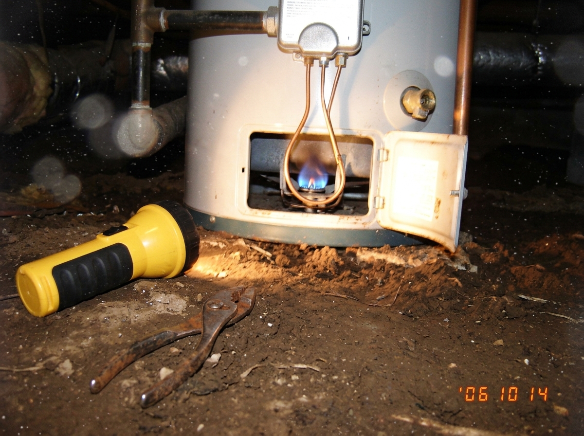 A close-up photo taken underneath a house, showing a yellow flashlight resting on a dirt floor next to a water heater's pilot light access panel. The panel is open, revealing a thin copper thermocouple tube and a small blue flame. The photo is slightly blurry and taken at an awkward angle, with dust