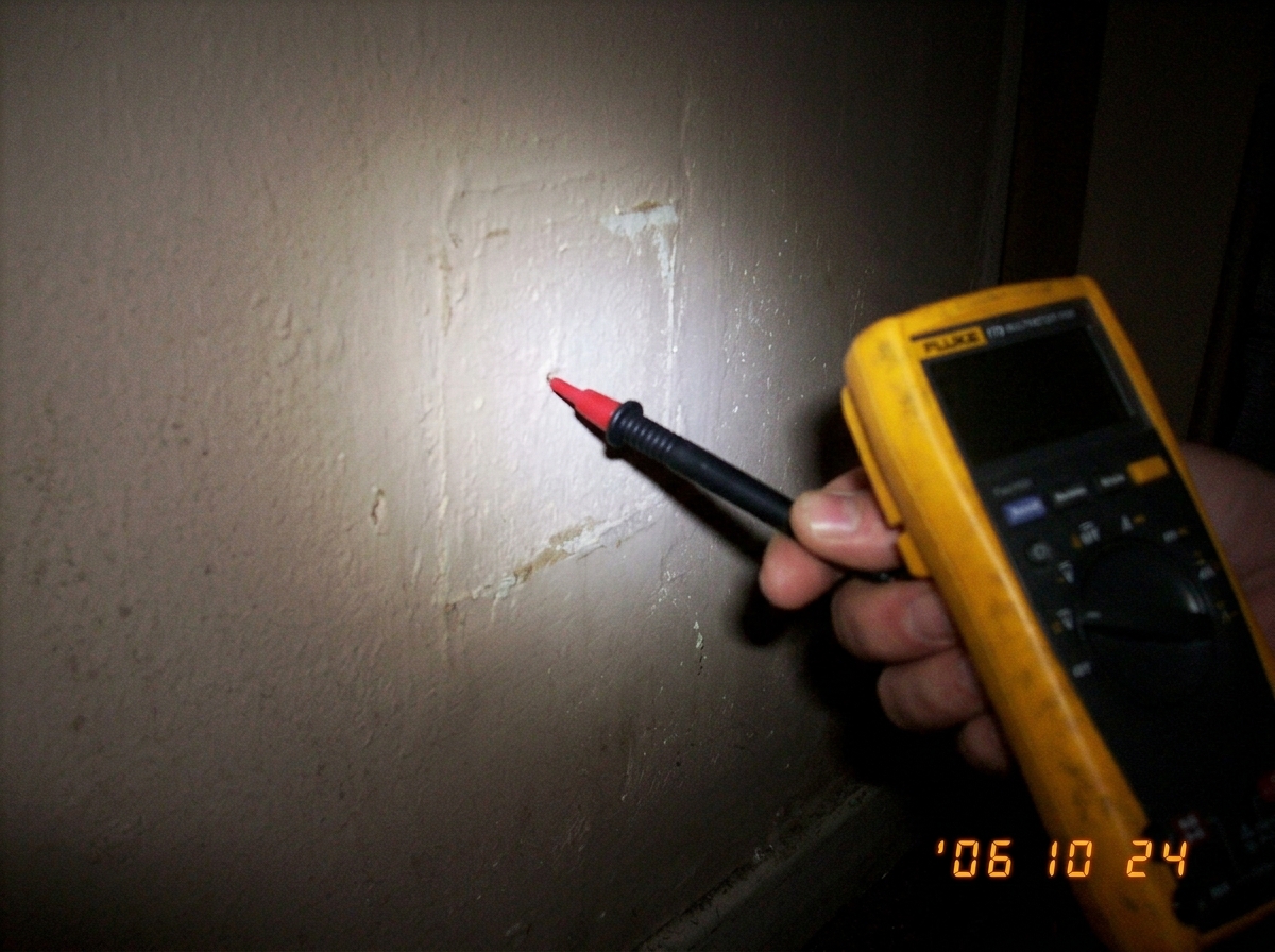 A close-up photo of a yellow Fluke multimeter probe pointing at a blank, empty wall where a socket should be. The lighting is dim.