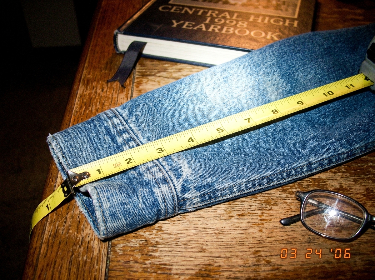 A very close-up, slightly blurry photo of a denim hoodie cuff laid flat on a wooden table. A yellow sewing tape measure is stretched across it, showing 47 threads per inch. In the background, an old high school yearbook is partially visible with a page marker ribbon. A pair of reading glasses rests 