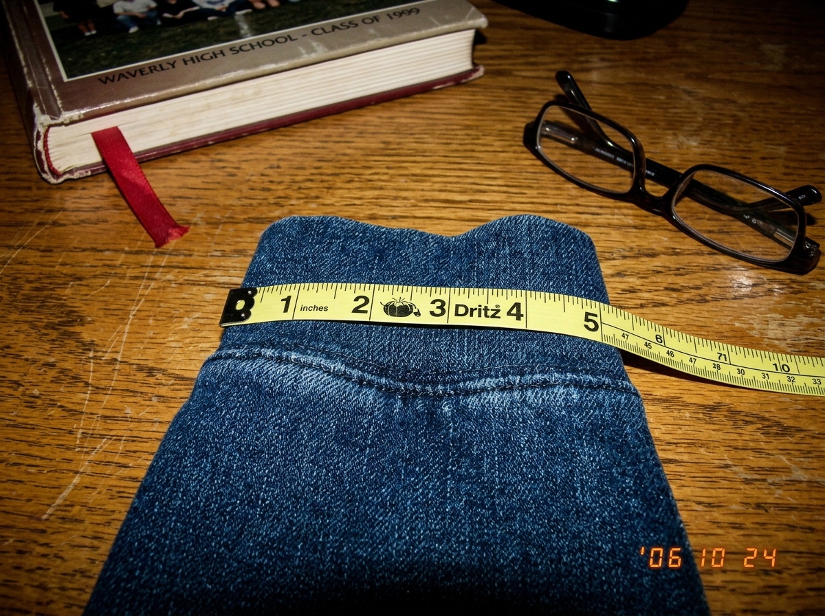 A close-up photo of a denim hoodie cuff laid flat on a wooden table. A yellow sewing tape measure is stretched across it, showing 47 threads per inch. In the background, an old high school yearbook is partially visible with a page marker ribbon. A pair of reading glasses rests on the table near the 