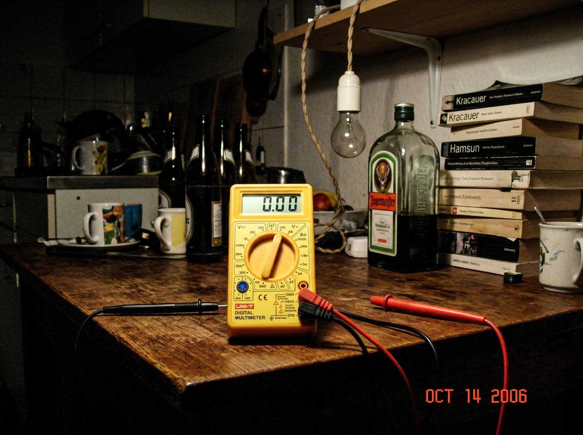 A photograph taken in a dim Berlin kitchen at night. A cheap digital multimeter sits on a wooden table, its probes touching nothing - just resting on the wood grain. The meter reads 0.00 but the backlight is on, casting a green glow on the surface. In the background, a bare lightbulb hangs from a tw
