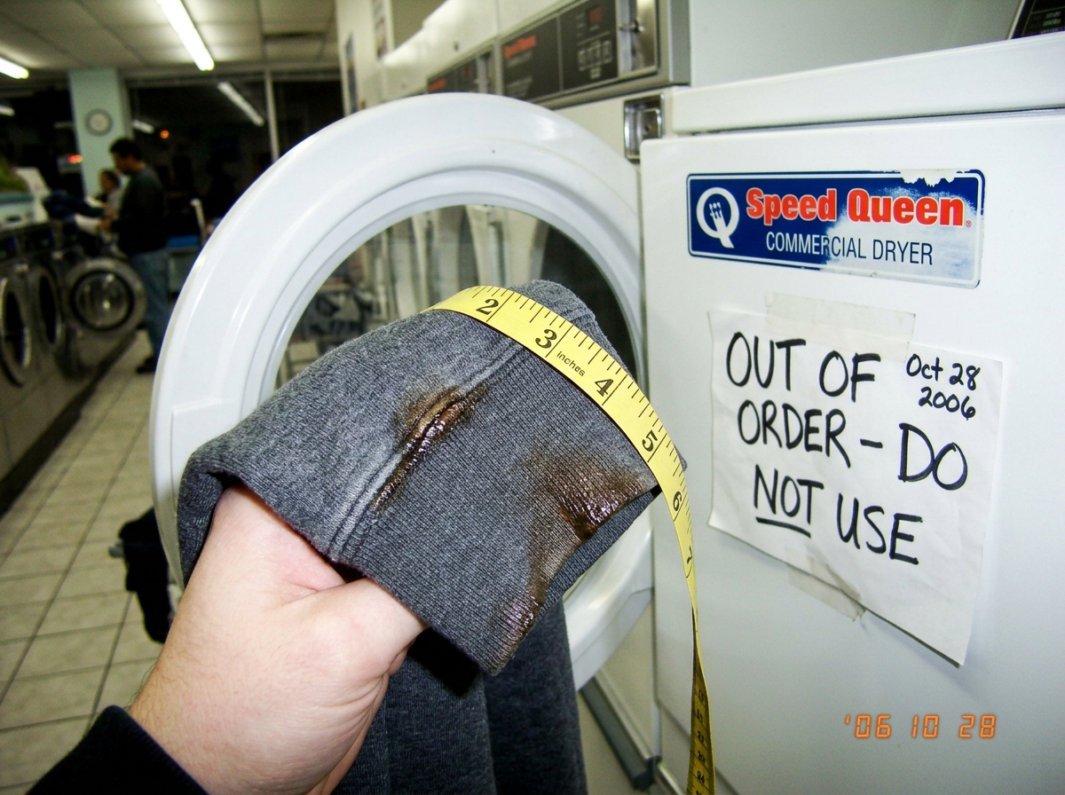 A person holding a burned grey hoodie cuff up against the open door of a white commercial dryer at a laundromat. The cuff shows a clear melt line where polyester threads have fused and discolored, with intact cotton weave visible on the other side of the thermal gradient. A yellow measuring tape is