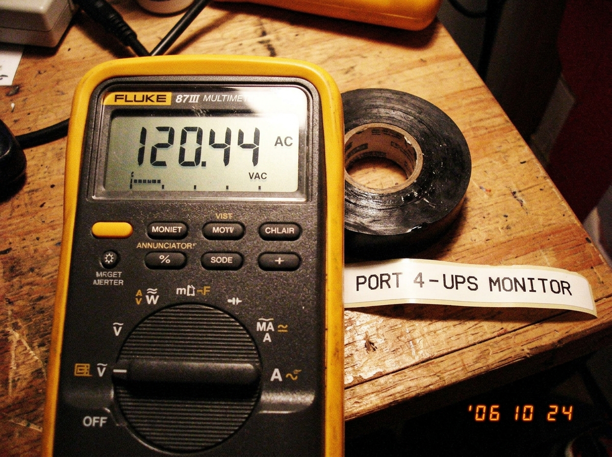 A close-up photo of a Fluke 87III multimeter sitting on a workbench next to a roll of electrical tape and a label maker printout reading 'PORT 4 - UPS MONITOR'. The meter display reads 120.4V AC.