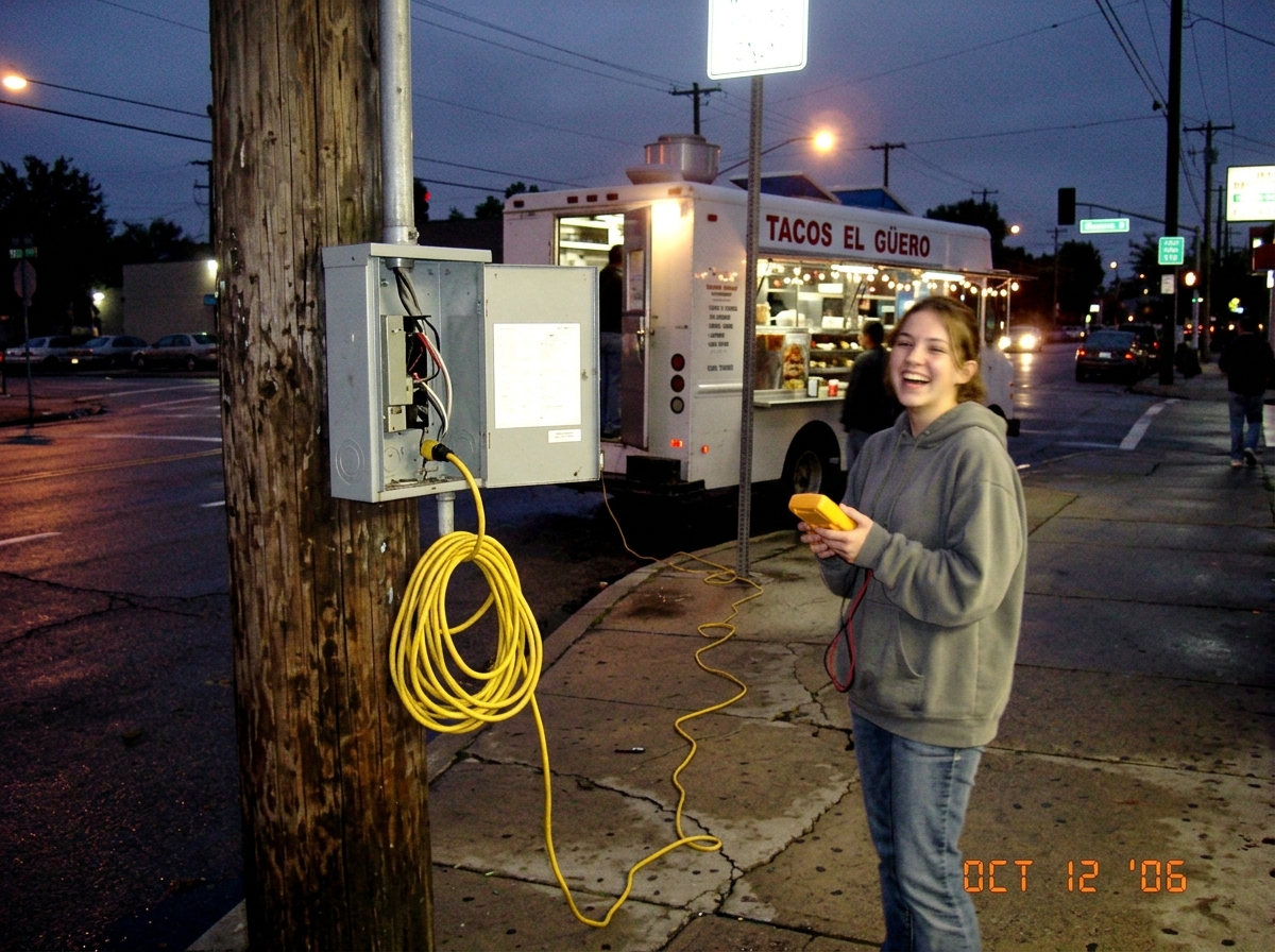 A photo taken at a street intersection at dusk. A grey junction box on a utility pole with the access panel slightly ajar. A yellow extension cord runs from the bottom of the box toward a sidewalk. In the background, a taco truck is visible with its lights on, and a girl in a grey hoodie is standing
