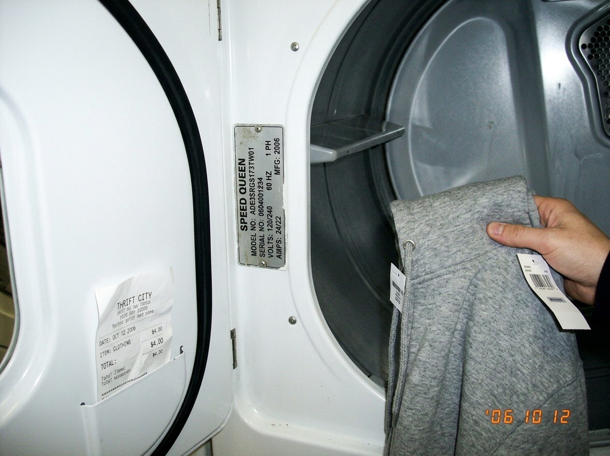 A photo of a Speed Queen dryer model plate inside an open dryer door at a laundromat. The plate reads 'ADE3SRGS173TW01' with serial number and electrical specs. Below it, a thrift store receipt for $4.00 is tucked into the door pocket, visible against the drum surface. A hand is partially visible ho