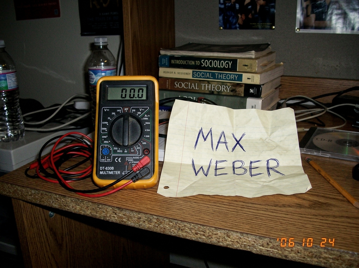 A low-res digital photo taken in a dimly lit dorm room showing a cheap digital multimeter sitting next to a crumpled piece of paper with 'MAX WEBER' scribbled on it