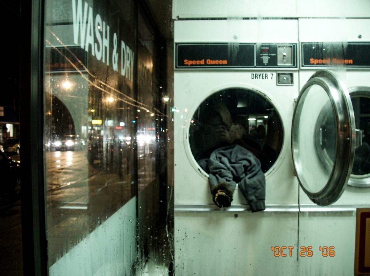 A blurry night photo taken through a laundromat window. Inside, a Speed Queen dryer is stopped with the door hanging open, a burned grey hoodie cuff visible against the drum. Reflected in the glass, a Euclid streetcar line passes by - the tram wires are visible as a faint double-line reflection cros