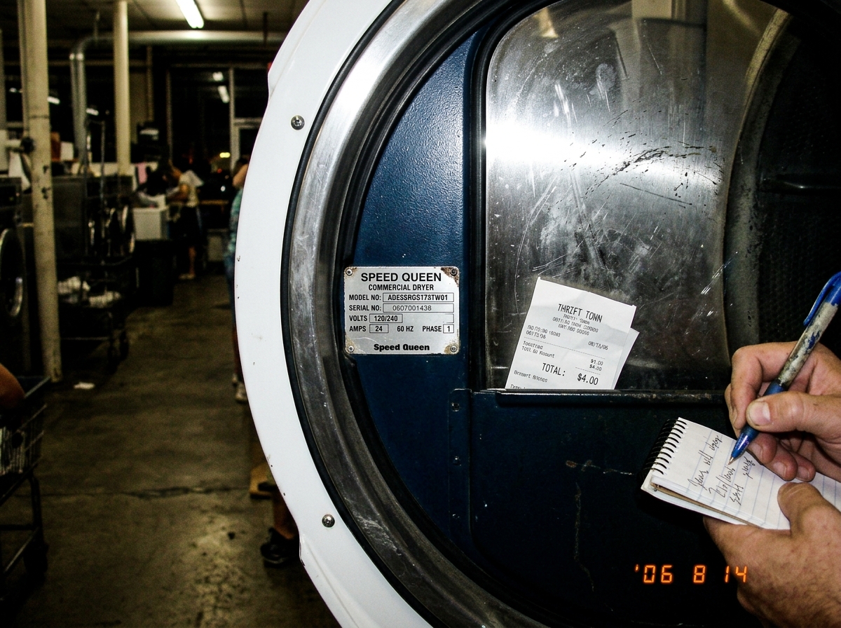 A close-up of a Speed Queen dryer model plate inside an open dryer door at a laundromat. The plate reads 'ADE3SRGS173TW01' with serial number and electrical specs. Below it, a thrift store receipt for $4.00 is tucked into the door pocket, visible against the drum surface. A hand is partially visible