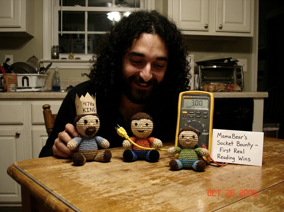 A photo showing three freshly crocheted Gabe-ables sitting on a kitchen table beside a Fluke 87V multimeter. One of the crocheted figures wears a tiny paper crown labeled '47Hz KING' and another holds a miniature yellow extension cord. A handwritten note beside them reads 'MamaBear's Socket Bounty -
