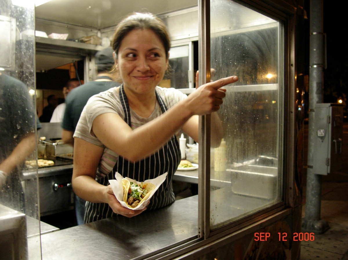 A blurry photo taken from inside a taco truck of a woman in her late 30s, slightly out of focus, holding a taco in one hand and pointing at the corner of the photo with the other hand, where the edge of a utility pole junction box is barely visible through the truck window. She has a knowing, secret