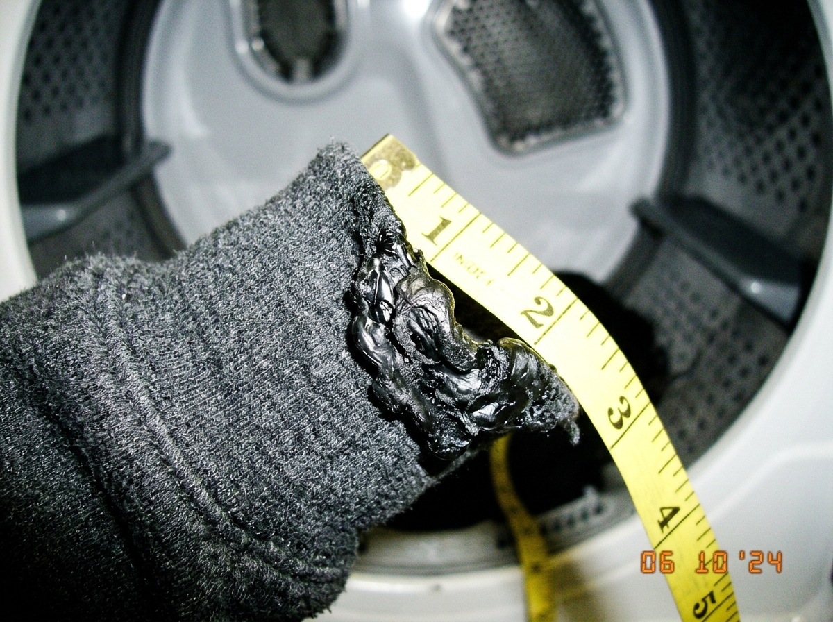 A close-up macro shot of a grey polyester hoodie cuff that has been melted into a hard, shiny plastic-like lump. A yellow tape measure is draped over the burnt fabric showing measurements near the edge. In the background, slightly out of focus, is a white commercial dryer drum interior.