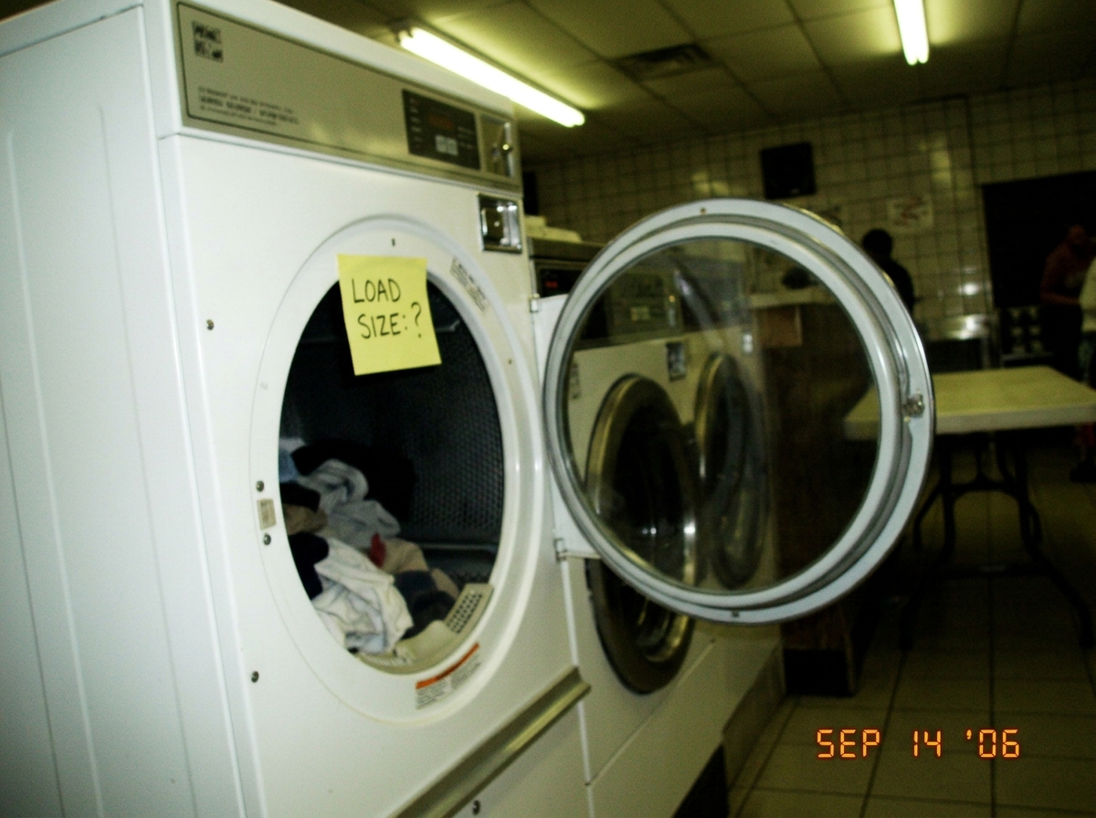 A blurry photo taken inside a laundromat at night, showing a white dryer with its door open. A handwritten sticky note on the drum reads 'LOAD SIZE: ?' in block capitals. The photo is slightly tilted and the fluorescent lights cast a greenish cast over everything.
