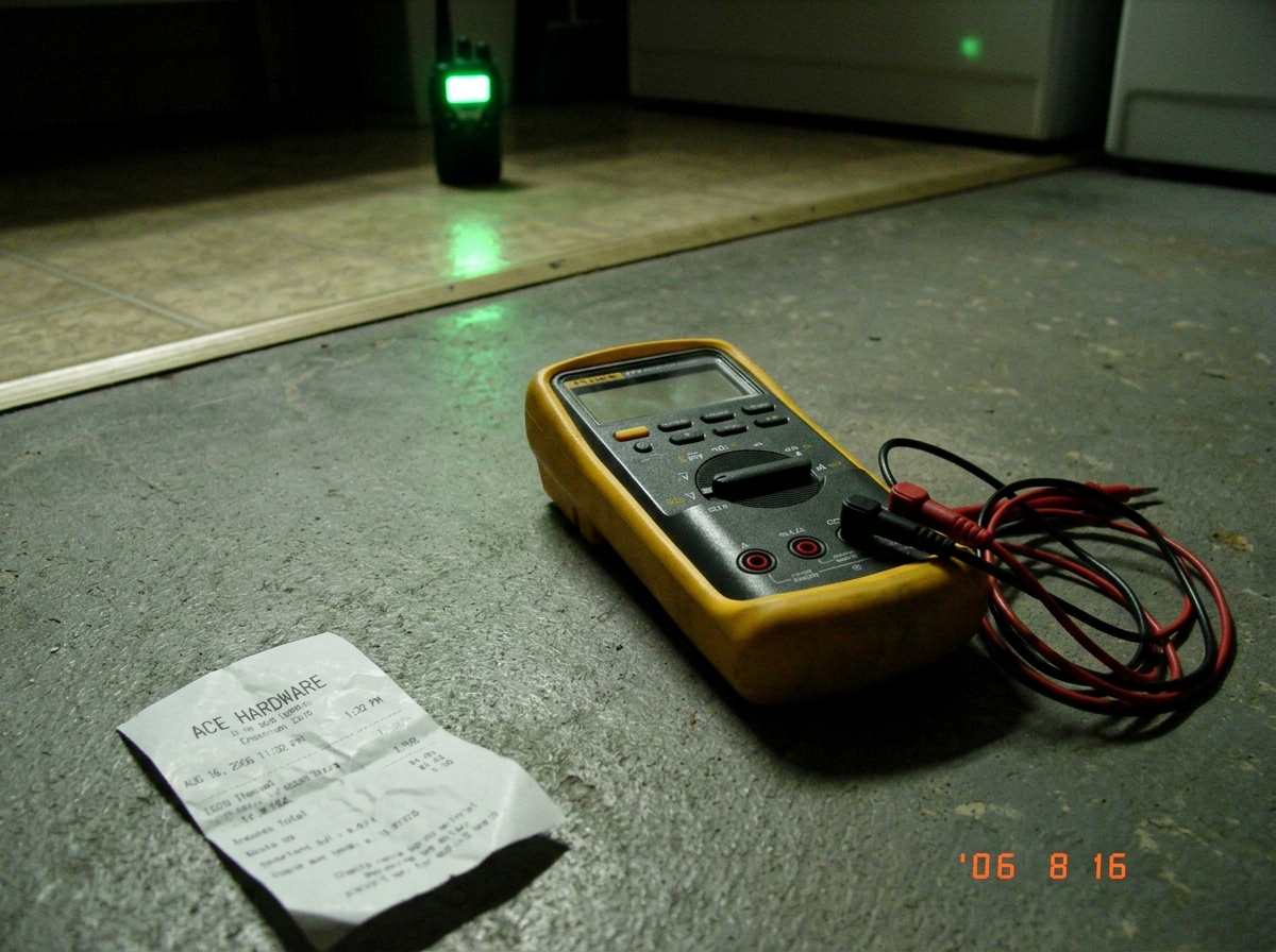 A slightly blurry flip phone photo of a Fluke 87V multimeter resting on a concrete laundry room floor. The leads are plugged into the meter but the screen is off. A crumpled Ace Hardware receipt in the corner shows a date of August 16, 2006 and a timestamp of 1:32 PM. In the background, a green LED