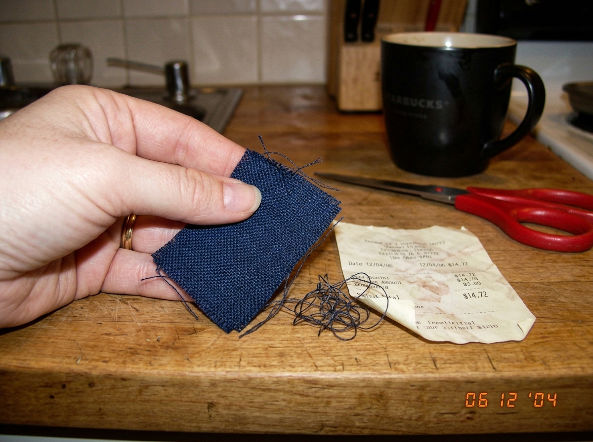 A blurry digital photo of a woman's hand holding a small piece of dark blue fabric with loose threads visible, next to a crumpled thermal receipt on a kitchen counter. The fabric has a visible weave pattern. A pair of scissors and a coffee mug are in the background.