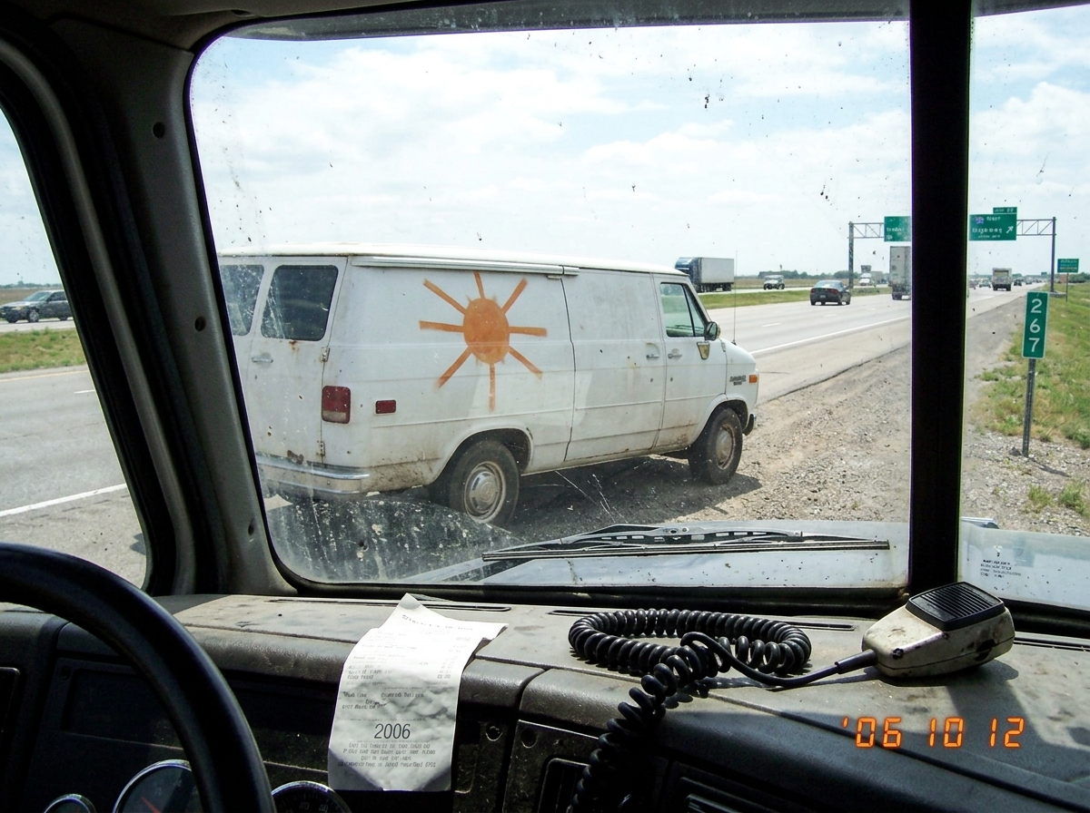 A photo taken from inside a semi truck cab showing a white mid-1990s Chevy van pulled over on a highway shoulder. The van's side door has a faded orange painted sun with eight straight rays radiating outward. The van is facing east. Through the truck's dusty windshield, mile marker 267 is visible. A