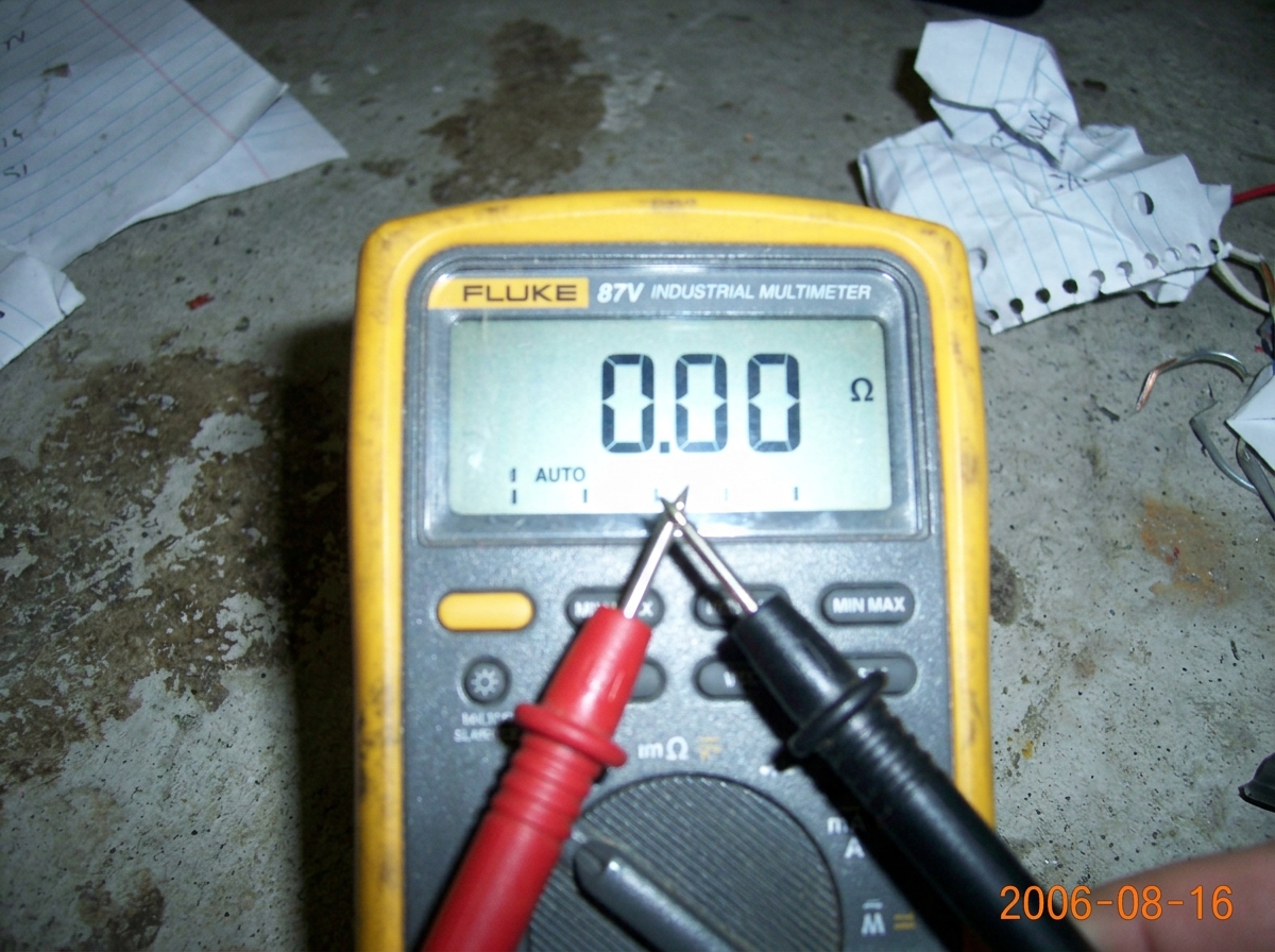 A close-up photo of a Fluke 87V multimeter display showing '0.00' while two bare probe tips touch each other against a blurred background of a concrete floor and crumpled paper. The date stamp on the camera reads 2006-08-16.