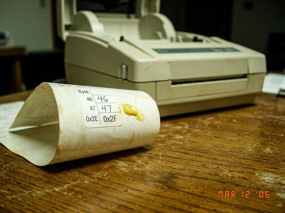 A thermal printer receipt curling on a stained wooden table. Three numbers visible on the serial sticker - 46, 47, and 0x2F - with a yellow mustard stain near byte 0x2E. A dead printer sits in the background, paper tray open.