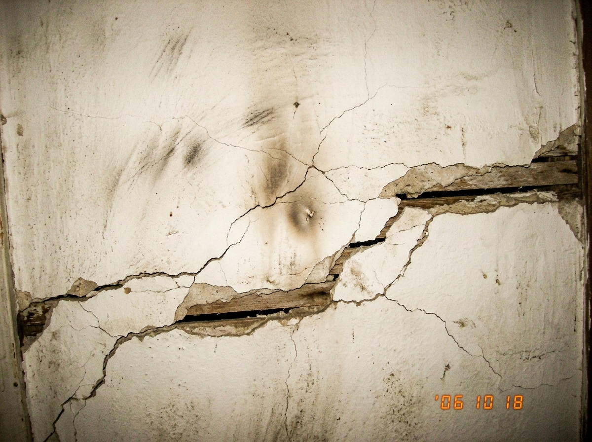 a close-up of a blank white wall showing faint scorch marks and hairline cracks where the plaster has separated from the lath, the texture of the old plaster visible