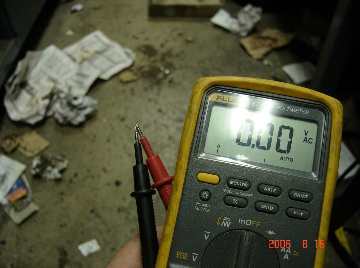 A close-up photo of a Fluke 87V multimeter display showing '0.00' while two bare probe tips touch each other against a blurred background of a concrete floor and crumpled paper. The date stamp on the camera reads 2006-08-16.