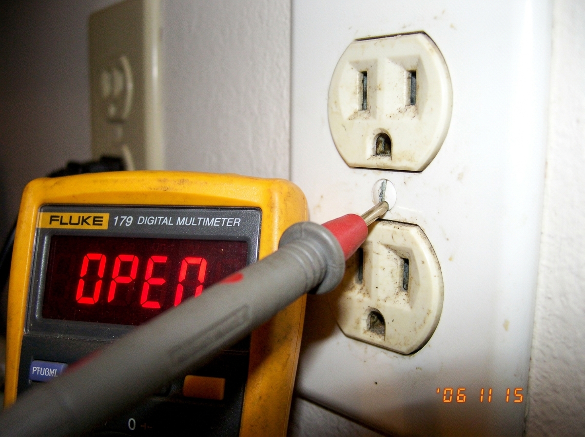 Close-up photo of a multimeter probe touching a dirty electrical outlet ground pin, with a digital display reading 'OPEN' in red digits