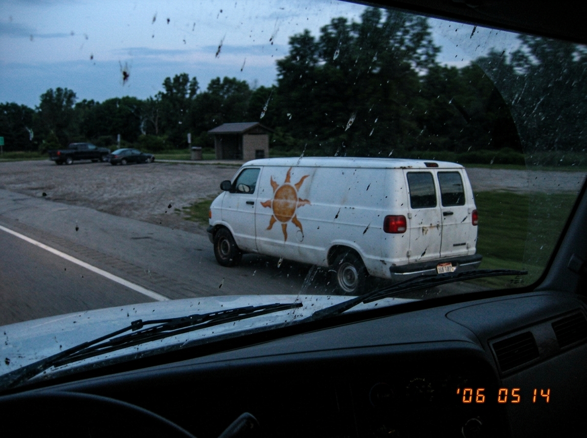 A blurry photo taken from a moving truck cab of a white church van pulled over at a highway rest stop, the side of the van visible showing a faded orange sun with eight rays painted near the sliding door, an Ohio license plate with 147 visible in the last three digits, early morning light, the photo