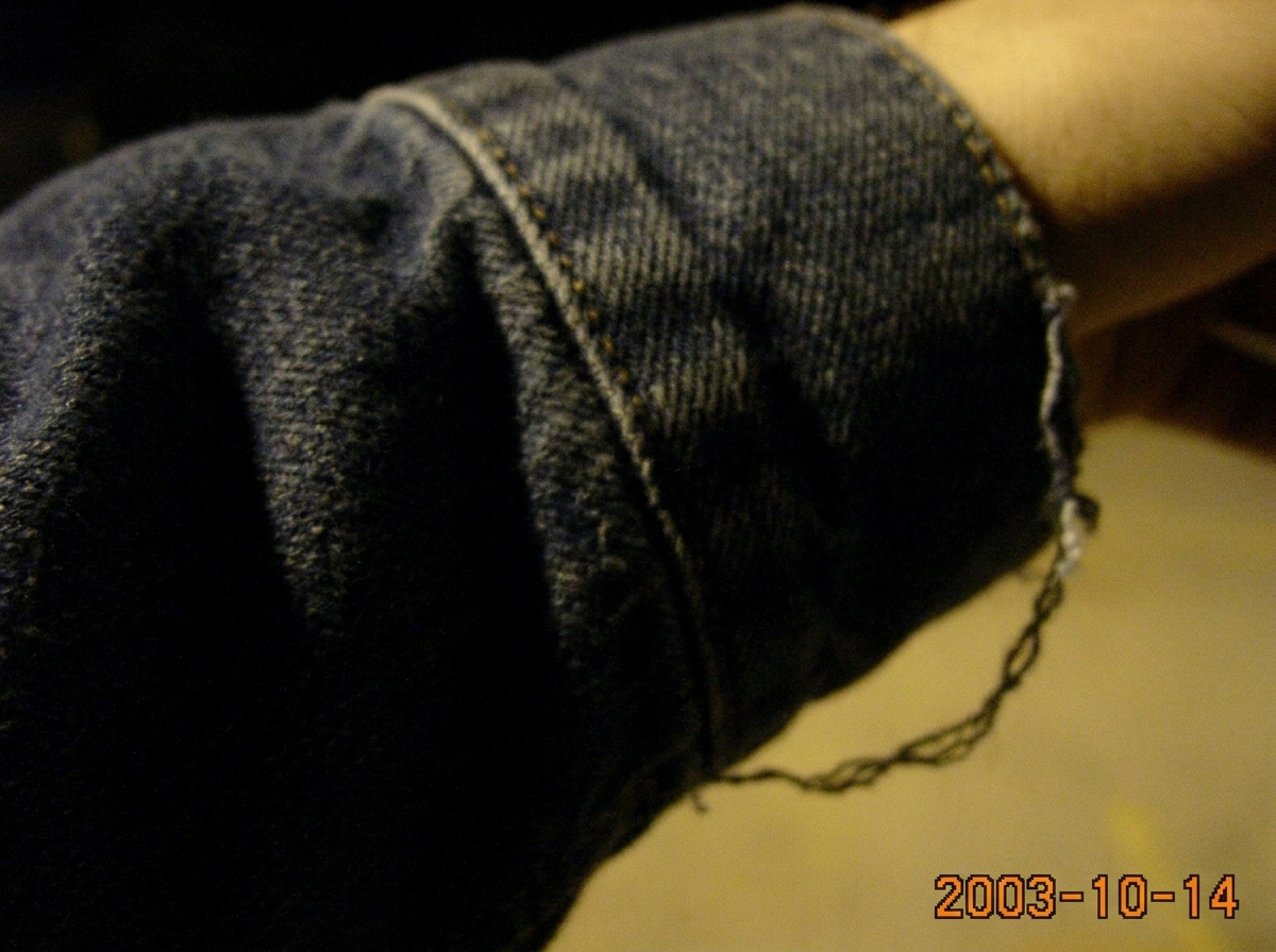 A blurry, heavily zoomed digital photo of a denim jacket cuff taken at night under a parking lot streetlight. The image is grainy and out of focus, but you can make out the weave of the denim fabric and what appears to be a loose thread hanging from the cuff edge. The timestamp in the corner reads '