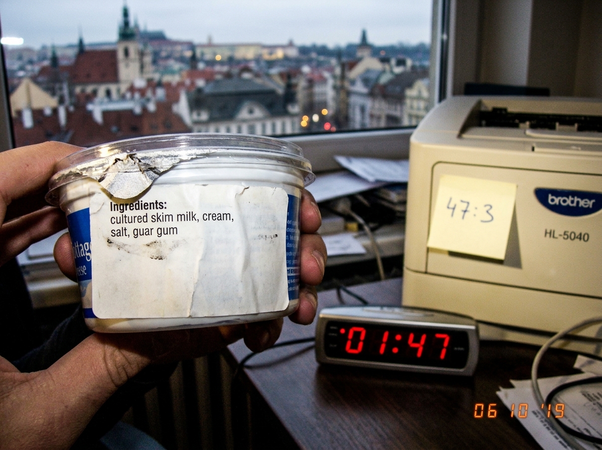 A macro photo of a cottage cheese container's ingredient label, held up against a window with a European city skyline visible through the glass. The label is torn at the corner and the ingredients list shows 'cultured skim milk, cream, salt, guar gum'. A digital clock on a nearby table reads 01:47. 