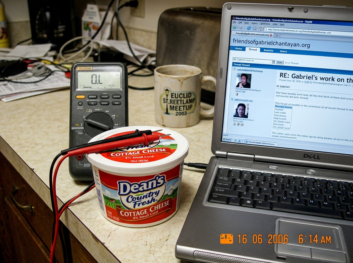 A close-up photo of a Dean's cottage cheese container sitting on a kitchen counter next to an open laptop. The laptop screen shows a forum thread from friendsofgabrielchantayan.org. A Fluke 87V multimeter sits nearby with probes resting on the cottage cheese lid. A coffee mug with a faded 2003 Eucli