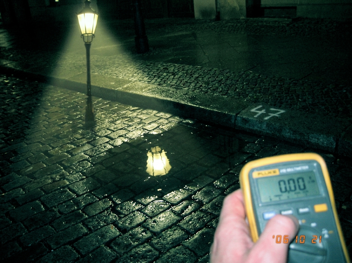 A nighttime photo of a wet Berlin street. A streetlamp casts a cone of yellow light onto the cobblestones, and in a puddle on the ground the reflection of the lamp is perfectly visible. A faint number 47 is spray-painted in white on the curb nearby. At the edge of the frame, someone's hand holds a F