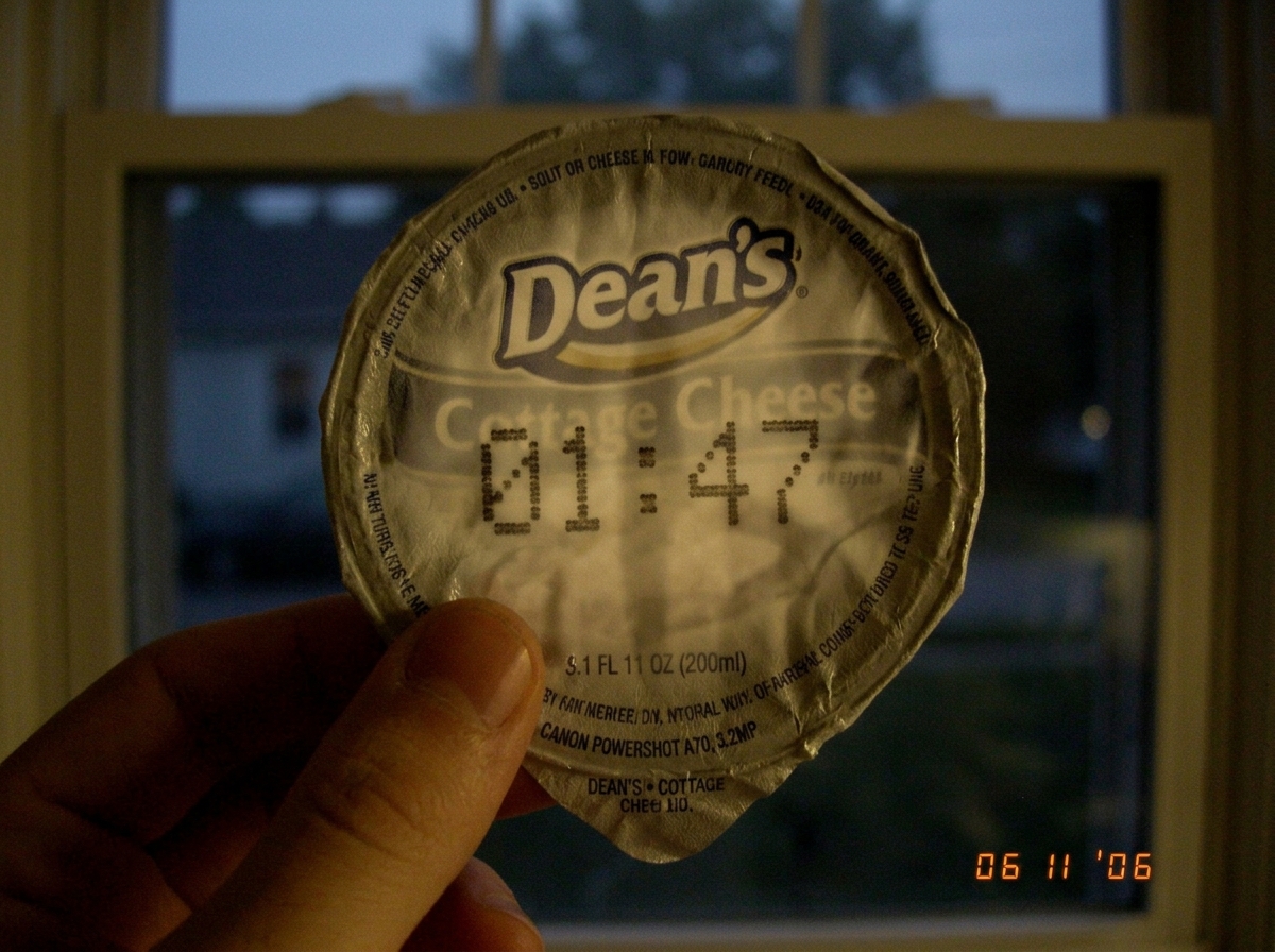 A dimly lit photograph of a hand holding a single, crumpled Dean's cottage cheese lid against the light of a closed window, highlighting the faint, concentric 01:47 timestamp shadow burned into the plastic, 2006 internet aesthetic