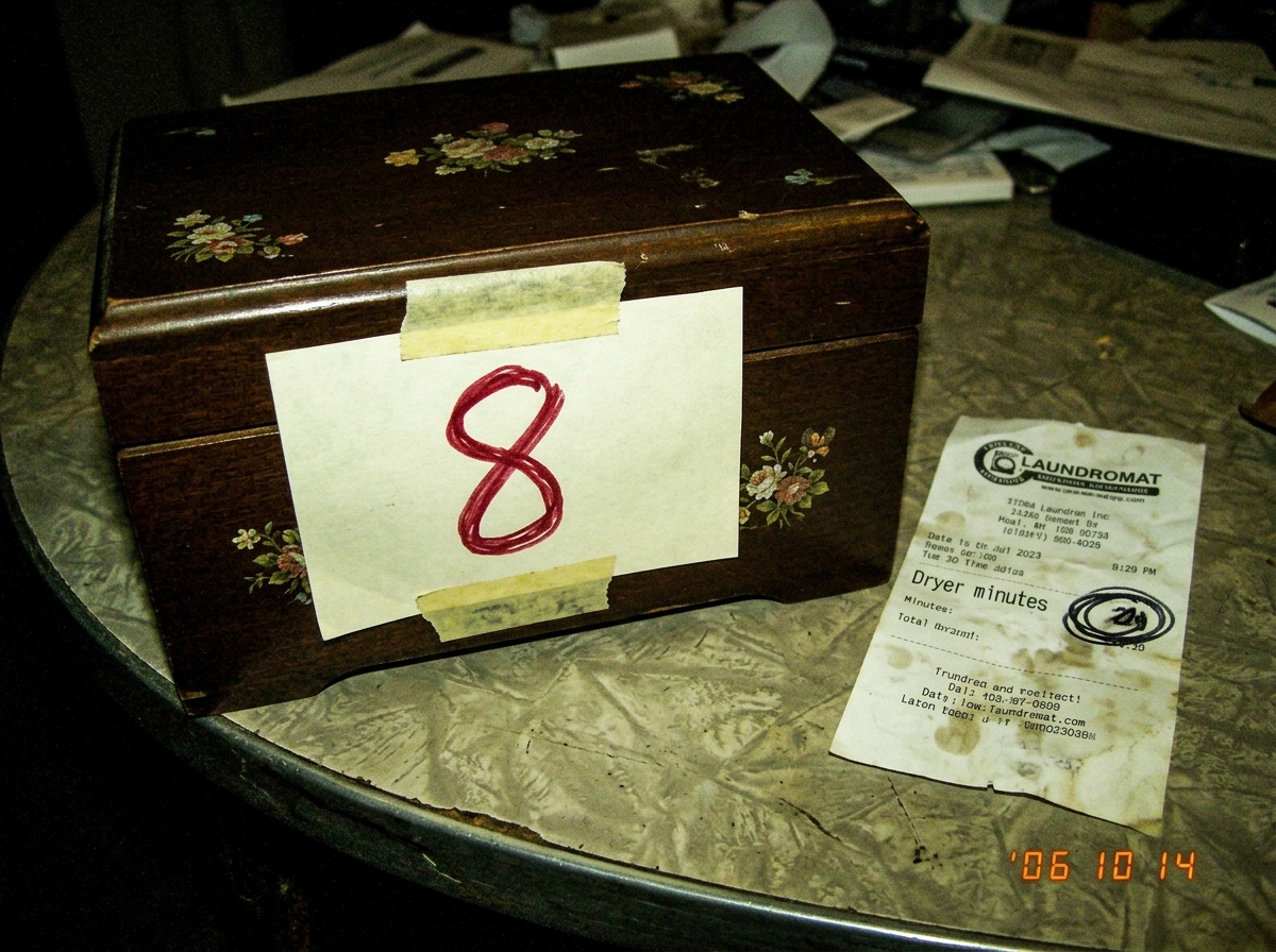 A grainy, poorly-lit photo of a sewing box with the number 8 crudely drawn on it in what looks like red marker on a piece of paper taped to it. The sewing box is the same one ClassOf04_Truther keeps posting - brown wooden box with floral decals. Beside it, a crumpled receipt from a laundromat with t