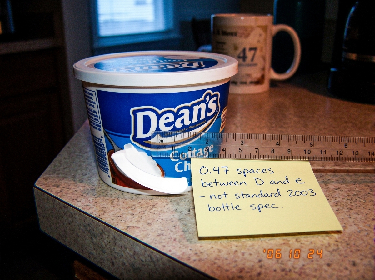 A close-up photograph of a Dean's cottage cheese container lid on a kitchen counter at dawn. The light from a window casts long shadows across the label. A small ruler is placed next to the word 'Dean's' showing the kerning measurement between the D and the e. A sticky note on the counter reads '0.4