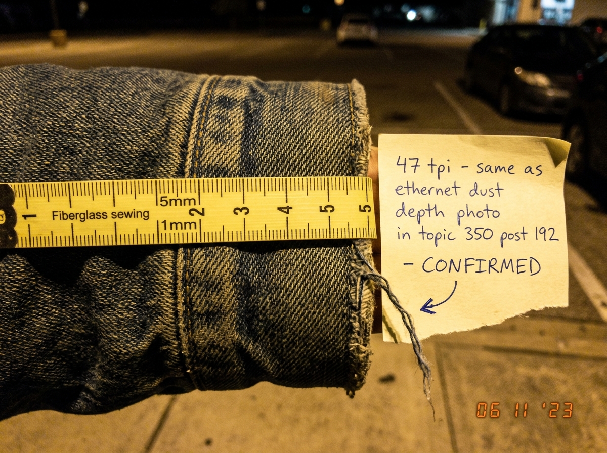 A macro close-up of a denim jacket cuff under parking lot lighting, with a literal sewing measuring tape laid across the fabric showing the weave count. A loose thread is visible hanging from the edge. A handwritten note on a torn piece of paper next to the cuff reads: '47 tpi - same as ethernet dus