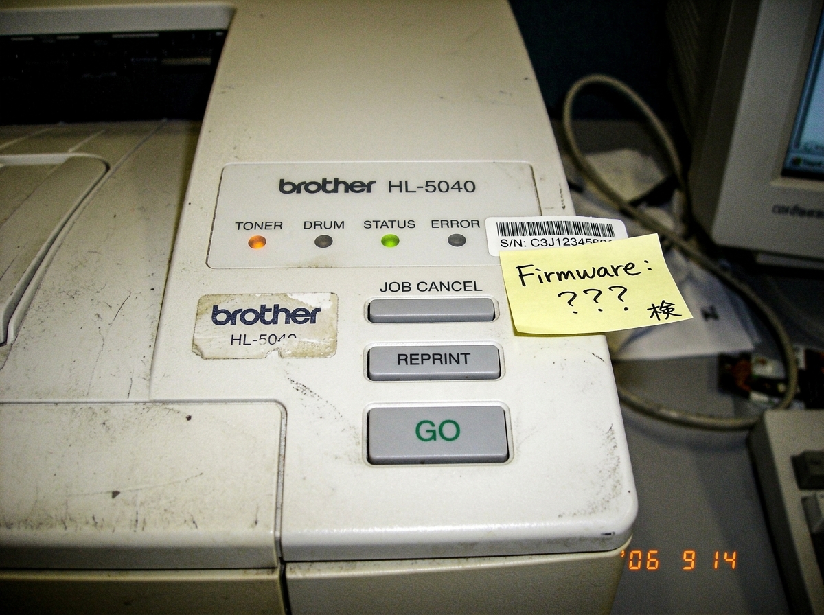 A close-up of a Brother HL-5040 printer's control panel with the serial number partially obscured by a small handwritten note saying 'Firmware: ???' and a tiny Japanese character '検' (check) next to it