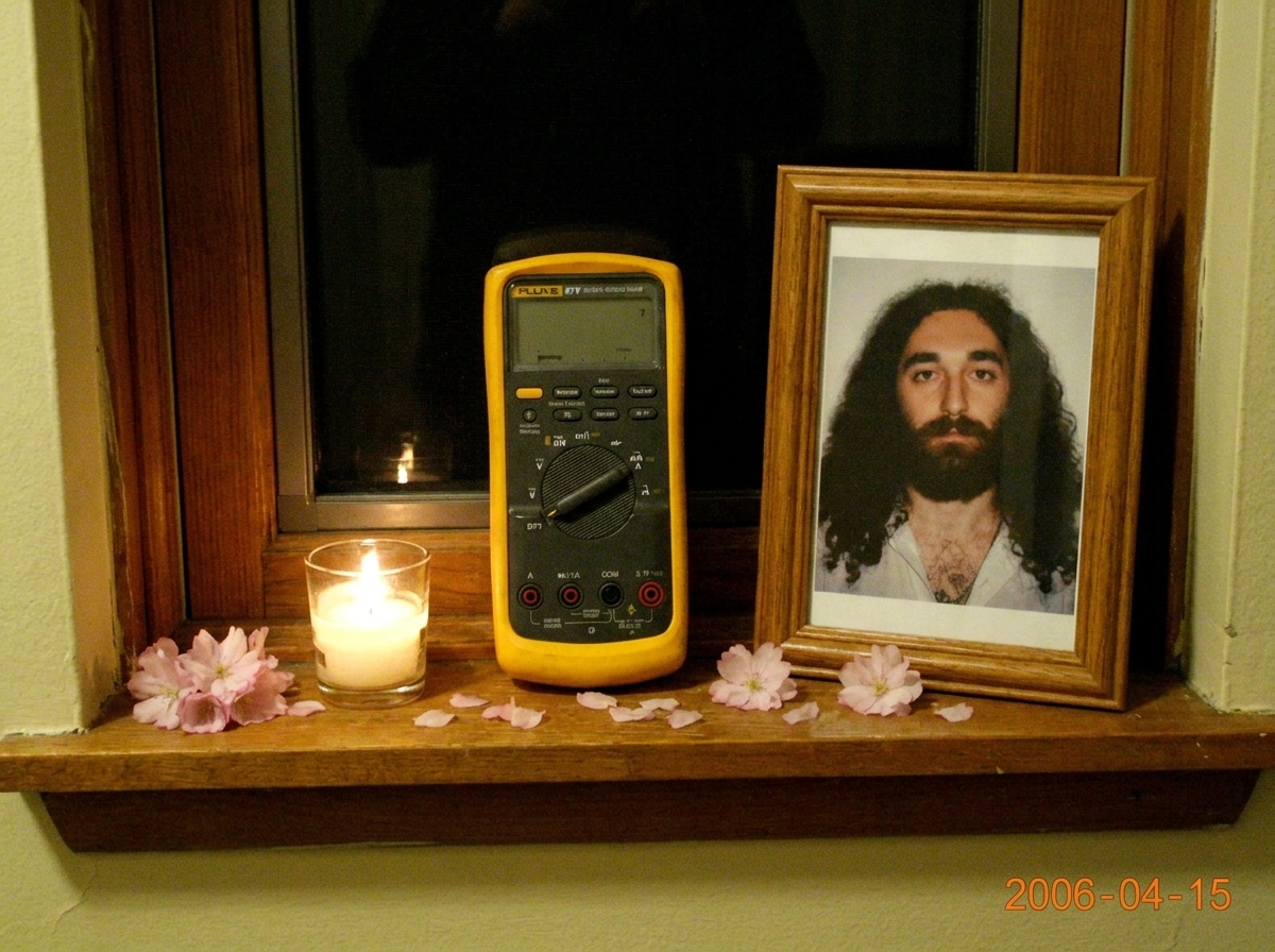 A small shrine on a windowsill with a Fluke 87V multimeter placed reverently next to a lit candle and a framed photo of Gabriel Chantayan, with cherry blossoms scattered around