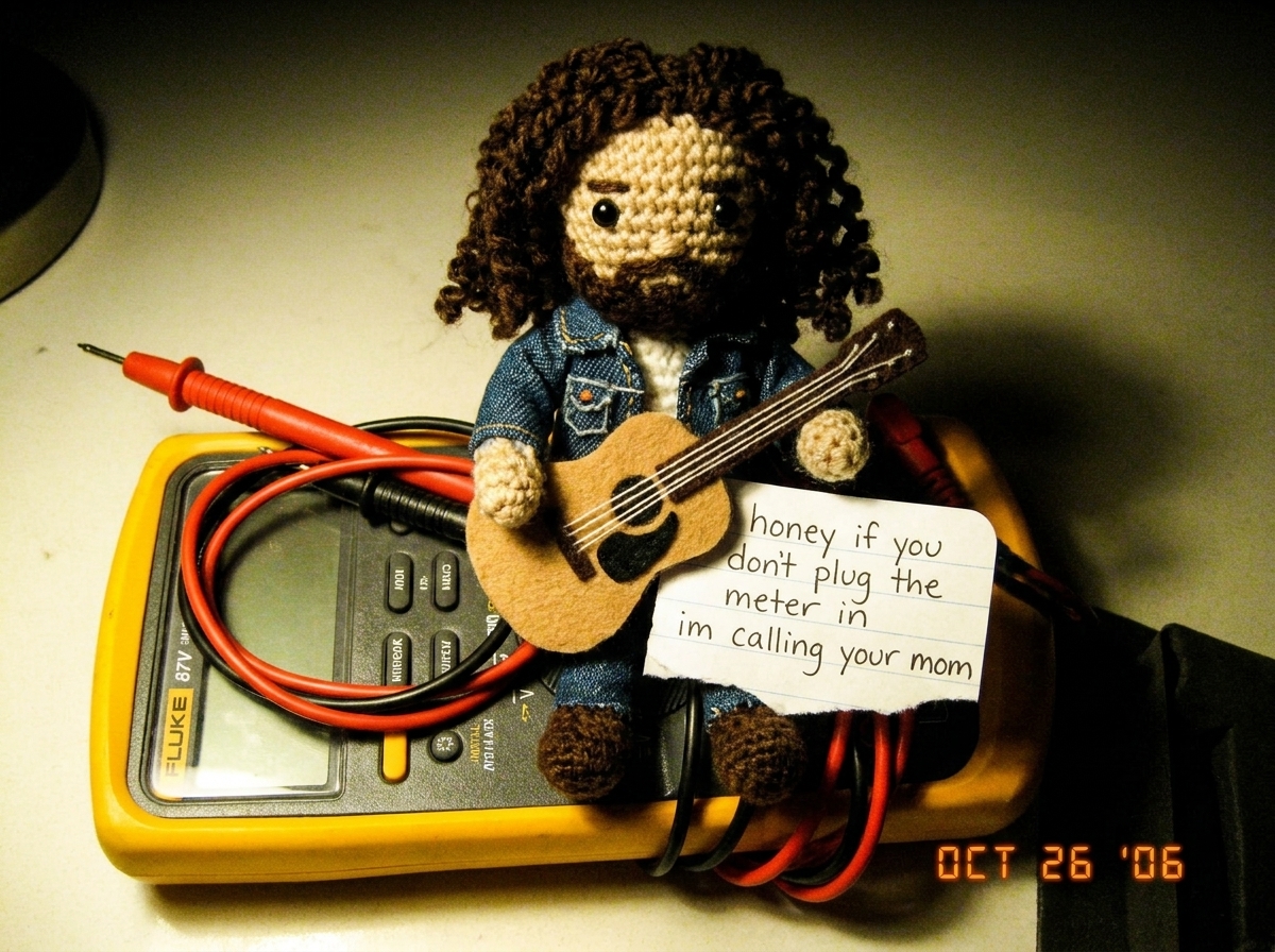 A small crocheted Gabe-able doll wearing a tiny denim jacket sitting on top of a Fluke 87V digital multimeter, the needle-probe leads coiled neatly beside it, with a hand-written note that reads 'honey if you dont plug the meter in im calling your mom' tucked under the Gabe-able's little felt guitar
