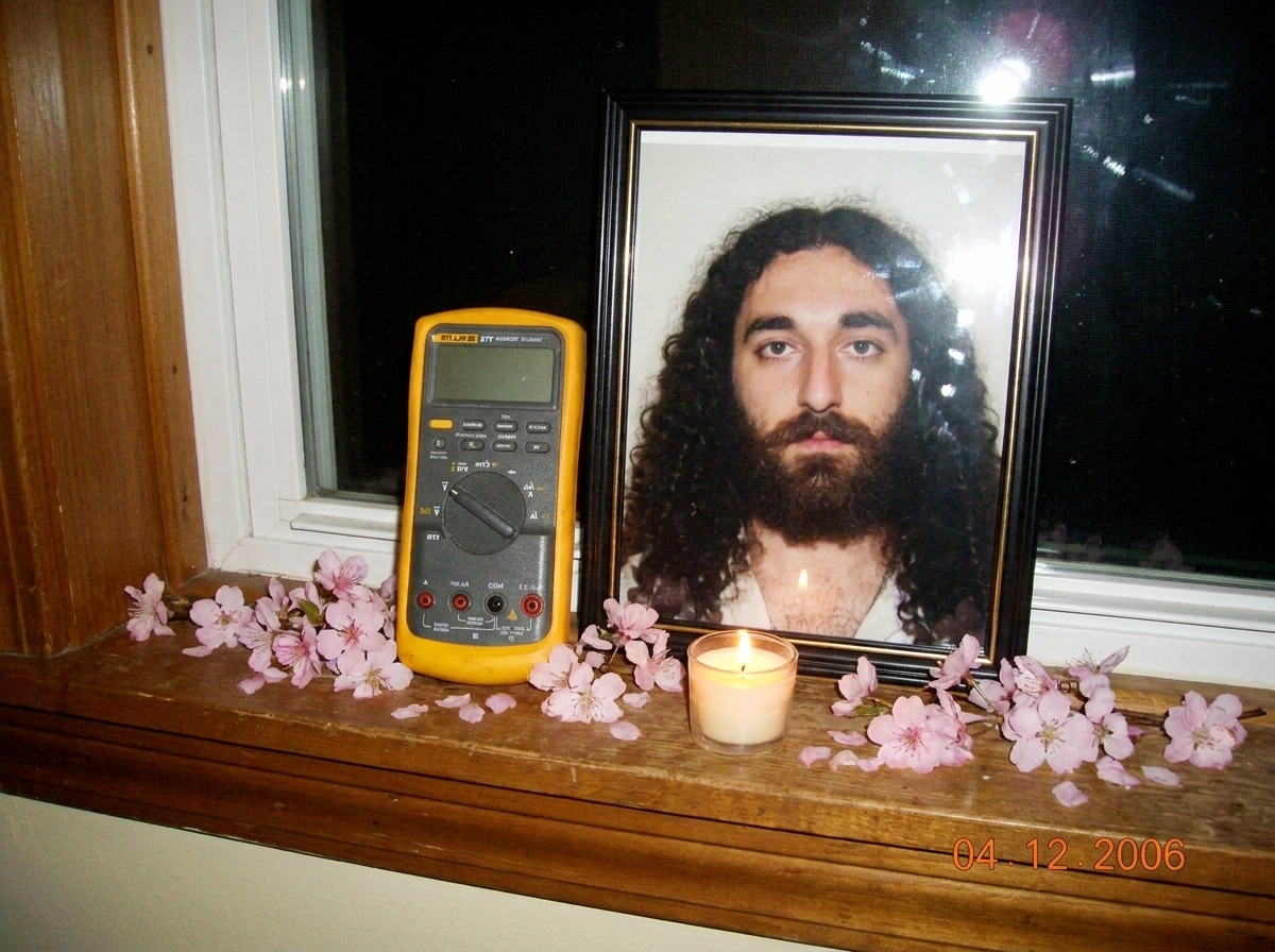 A small shrine on a windowsill with a Fluke 87V multimeter placed reverently next to a lit candle and a framed photo of Gabriel Chantayan, with cherry blossoms scattered around
